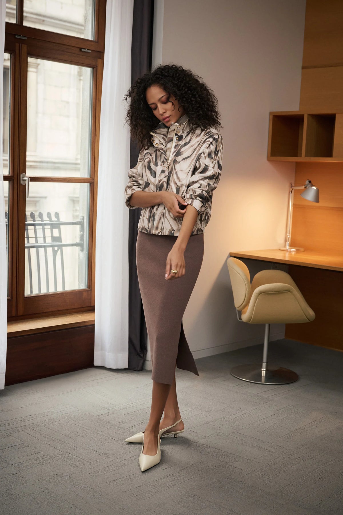A woman stands in a modern office setting wearing a patterned long-sleeve blouse, a JOSEPH RIBKOFF SWEATER KNITT MIDI SKIRT IN MINK 232967F26, and cream pointed-toe flats. The background features a desk, a chair, and a window with curtains.