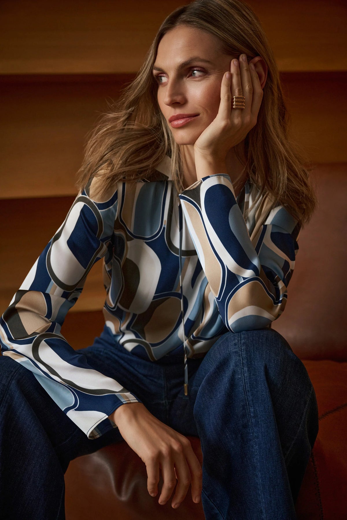 Woman wearing a patterned blouse sitting on a couch. Joseph Ribkoff Geometric V-Neck Collar Blouse in Off-White/Multi, style 263071 