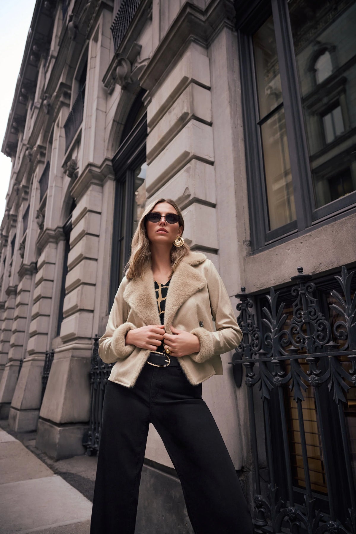 Woman in a beige coat and sunglasses standing in front of an architectural building. Full-length styled outfit featuring Joseph Ribkoff Faux Fur Trim Moto Jacket in Soft Taupe, style 263909