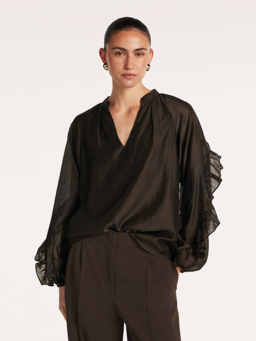 A model wearing beaumont amanda blouse dark brown with a deep V-neckline and ruffled cuffs, paired with dark brown trousers, against a plain off-white background.