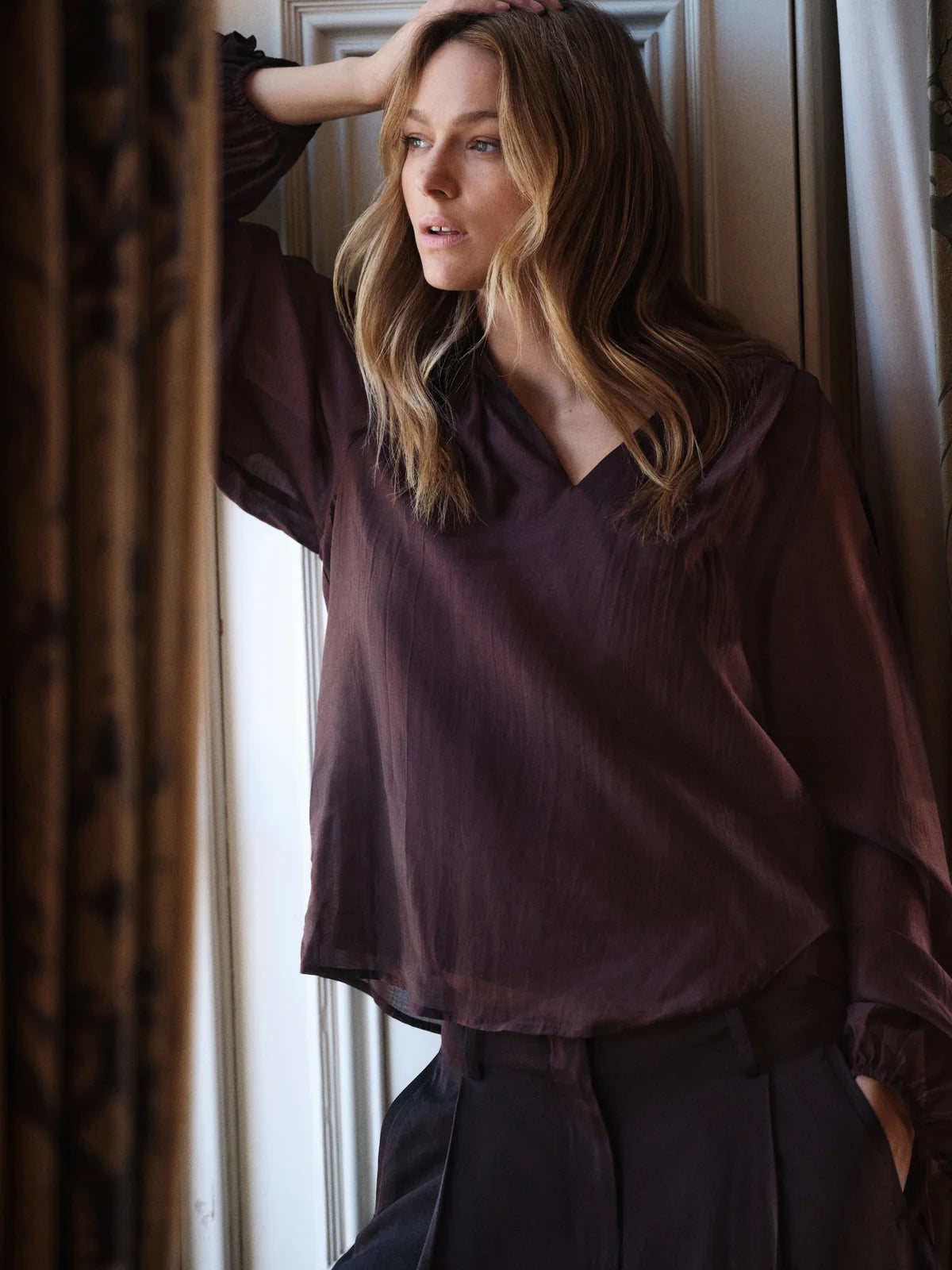 A woman wearing amanda blouse dark plum  and dark trousers, leaning against a window with her hand on her head, framed by patterned curtains.