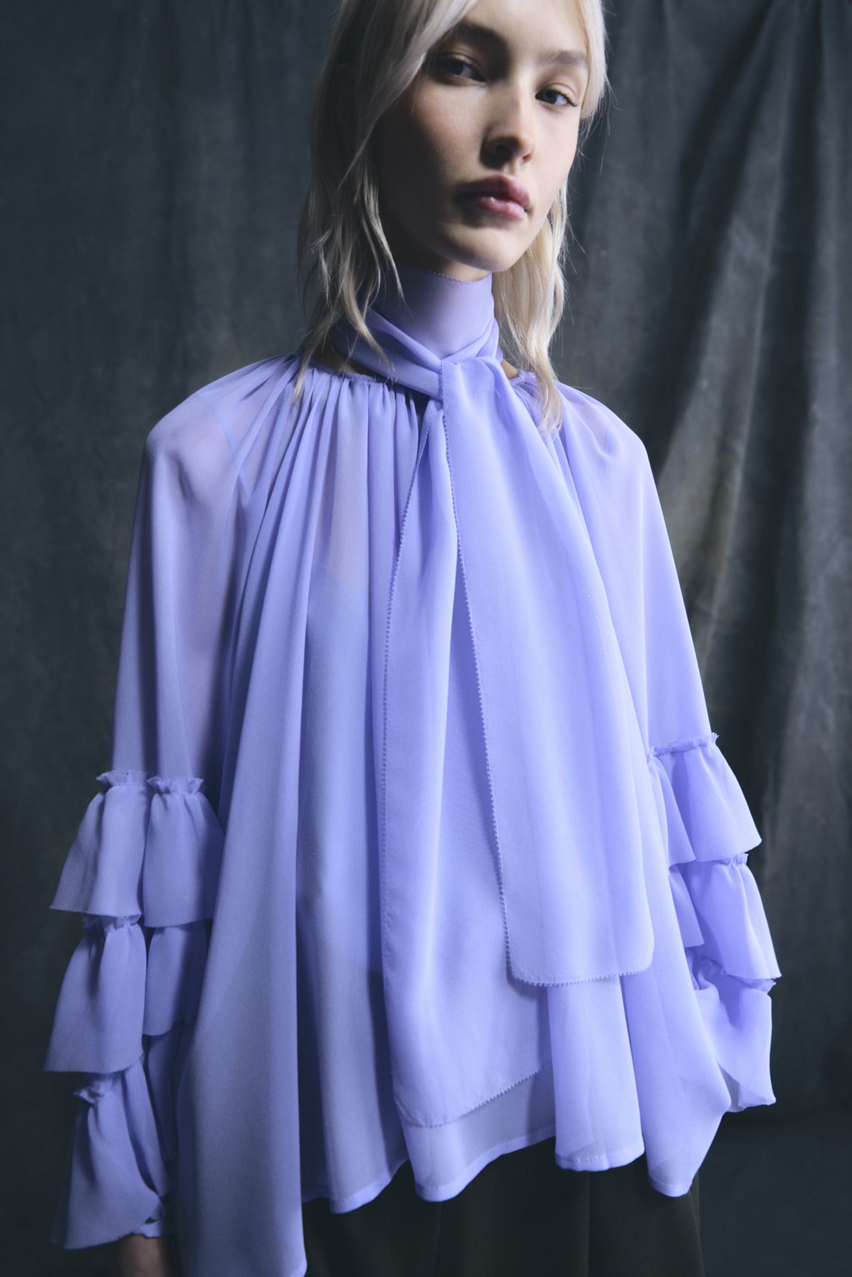 Woman wearing a Psophia thystle blouse light purple blouse with ruffled layers against a dark background